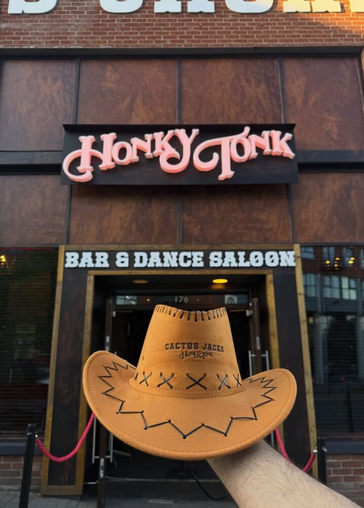 A tan suede Sonison cowboy hat printed with the Cactus Jack's Honky Tonk logo, held up outside the Honky Tonk Bar and Dance Saloon